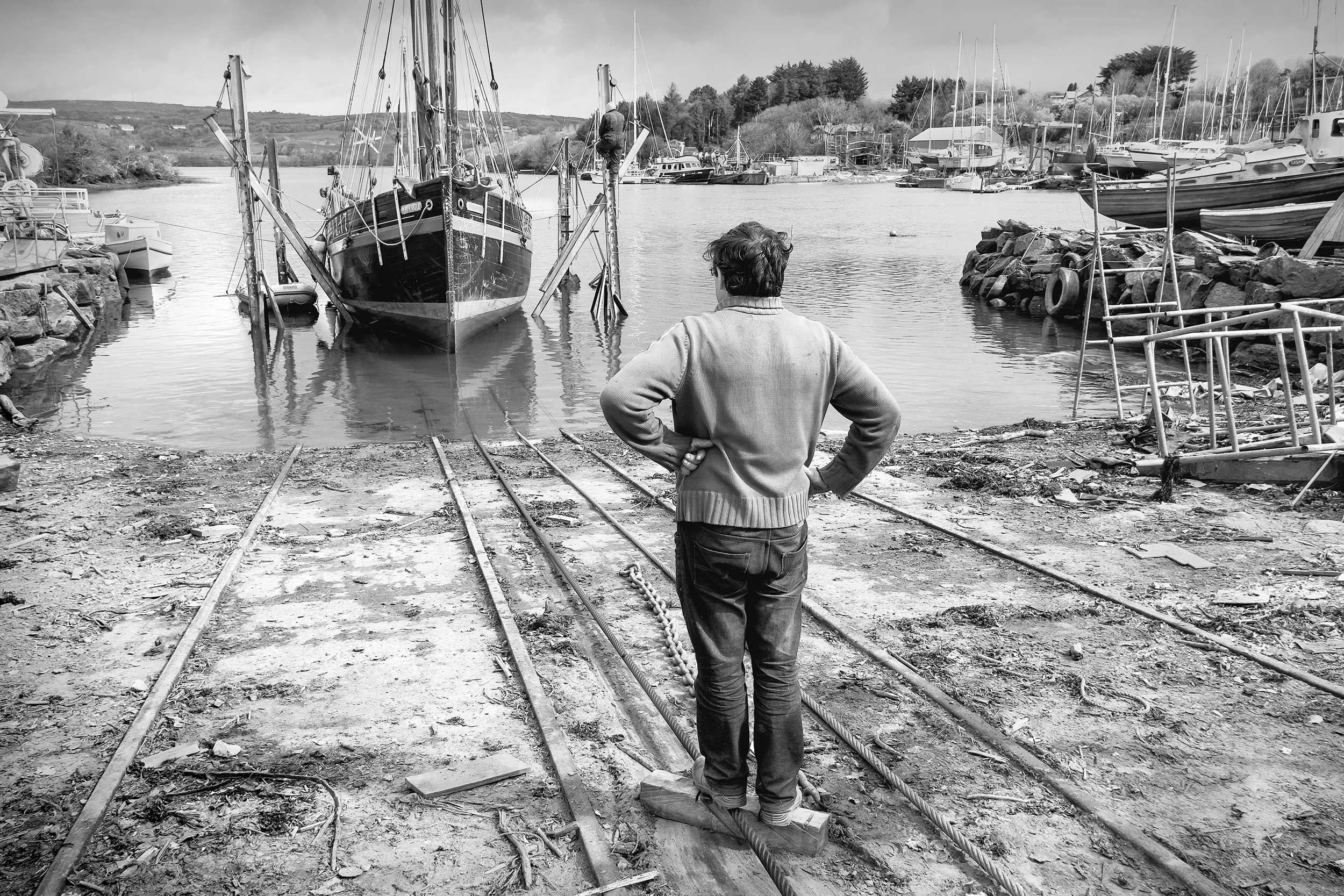 Black and white photo of a man standing with his hands on his hips, looking at a ship undergoing maintenance.