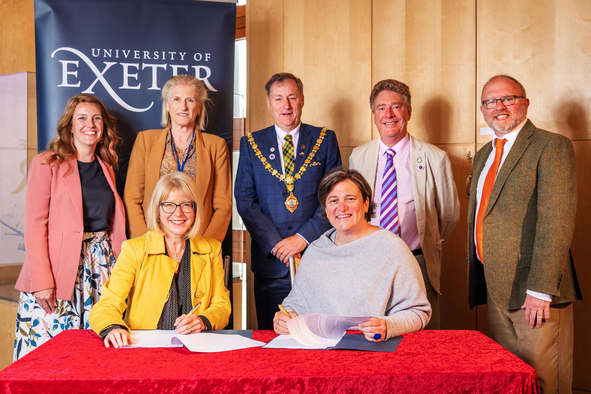 Seven people - four women and three men - pose for a photograph in front of a 'University of Exeter' banner. Two of the women are seated at a table with a red throw over it, signing two documents.