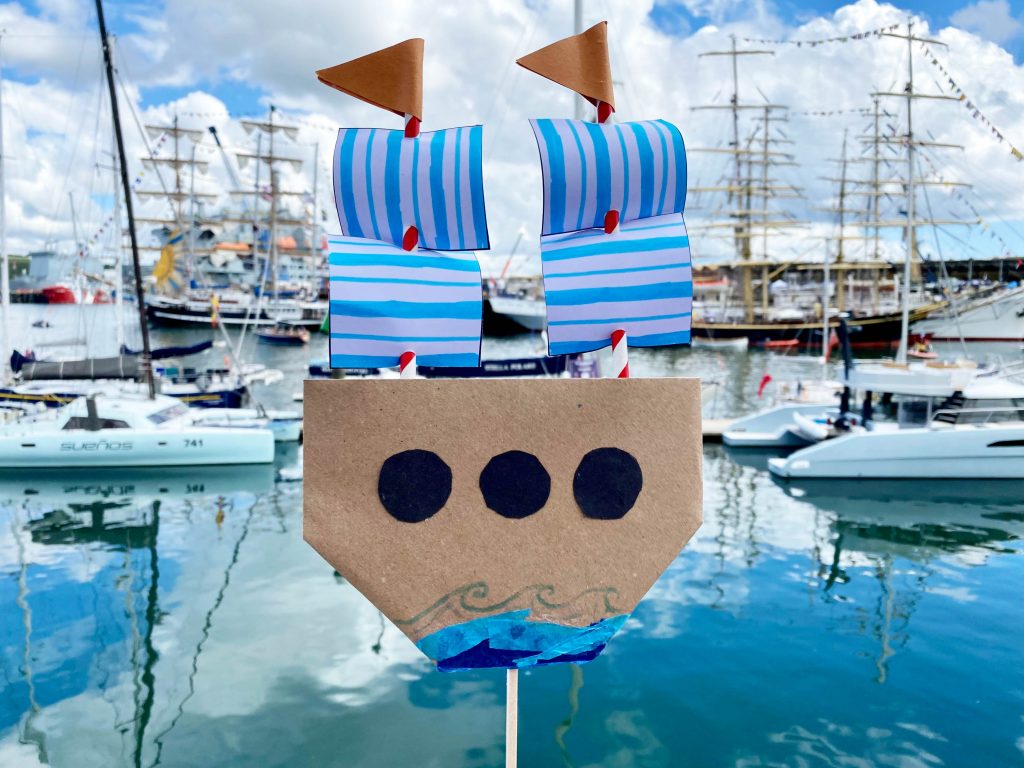 A photo of a handmade tall ship made from cardboard, paper and straws, with two Tall Ships in Falmouth Harbour in the background. Photographed on a bright, sunny day.