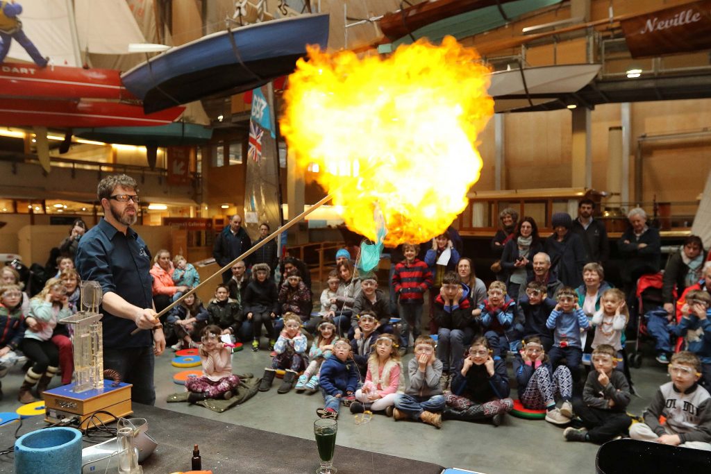 Standing in front of a crowd of young children and their parents, a man carries out a science experiment that results in a ball of fire.