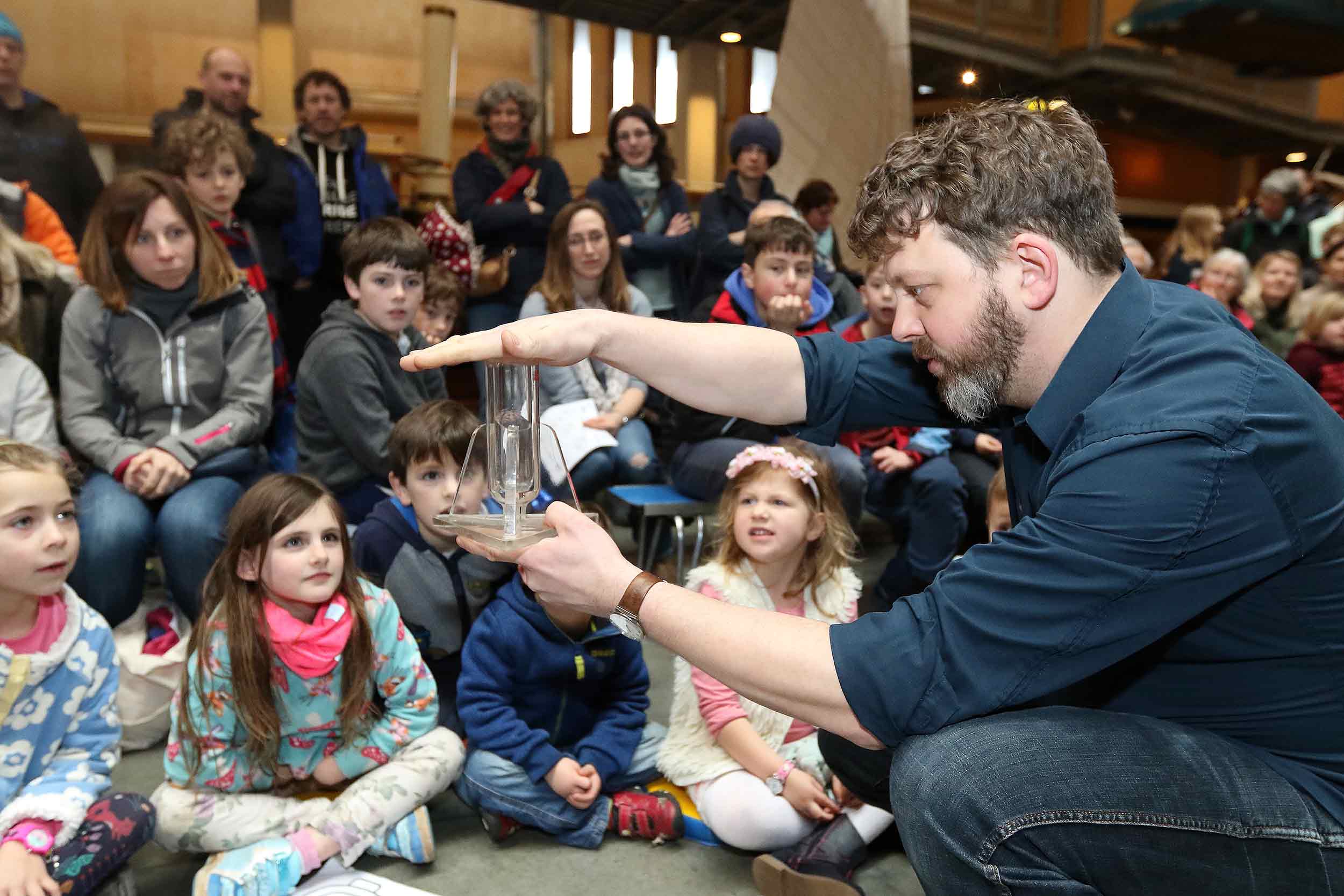 A man crouches down in front of a group of children to conduct a science demonstration.