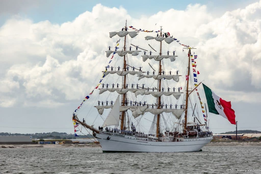 A photo of the Mexican tall ship Cuauhtemoc. A large white sailing ship flying the Mexican flag, pictured on a sunny and cloudy day.