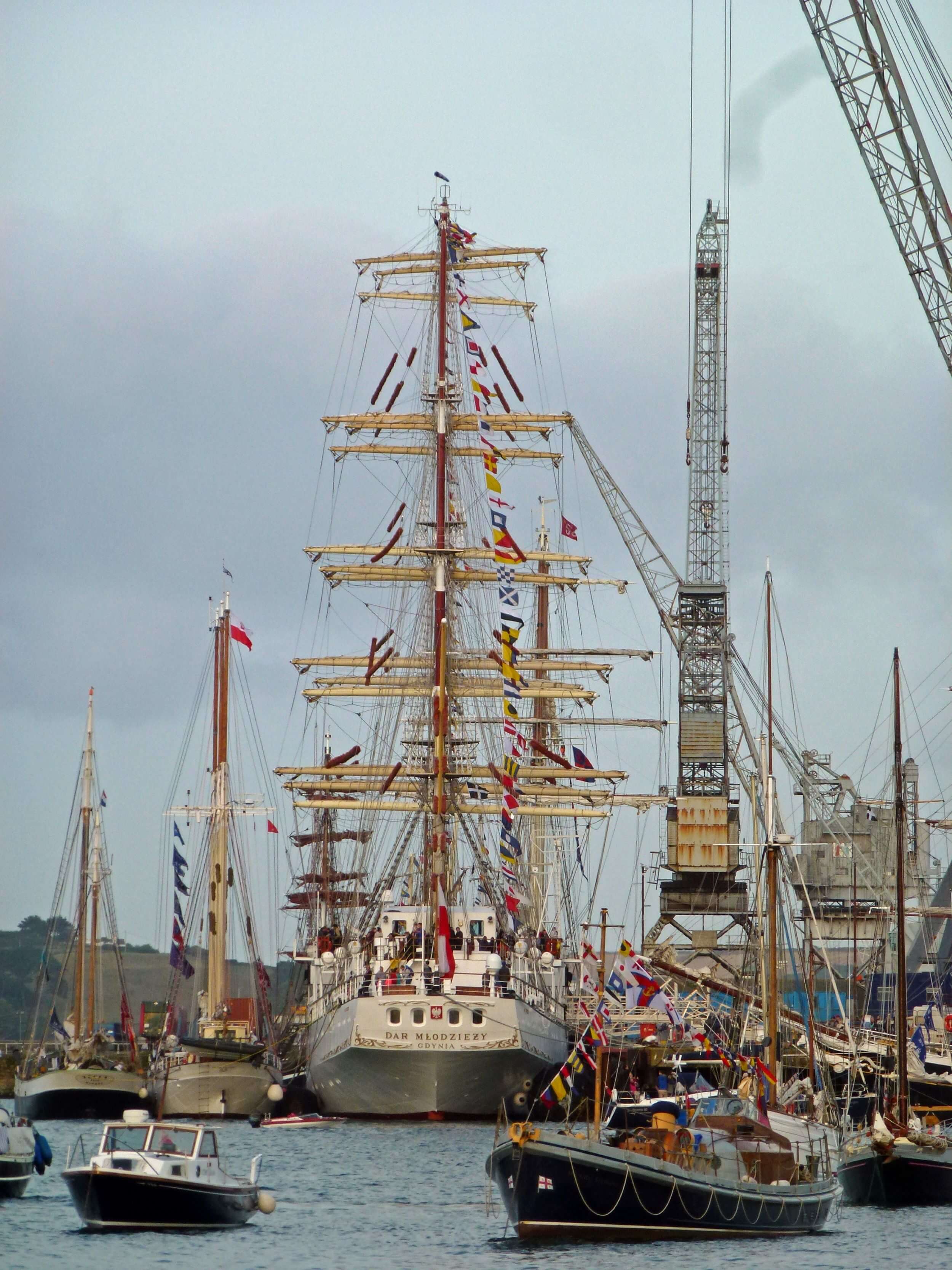 A photo of the Polish Tall Ship Dar Mlodziezyat A&P Falmouth in 2014. Taken during the regatta, the ship is surrounded by a variety of boats, including other tall ships, a historic lifeboat, and small sailing vessels. It is an overcast day.