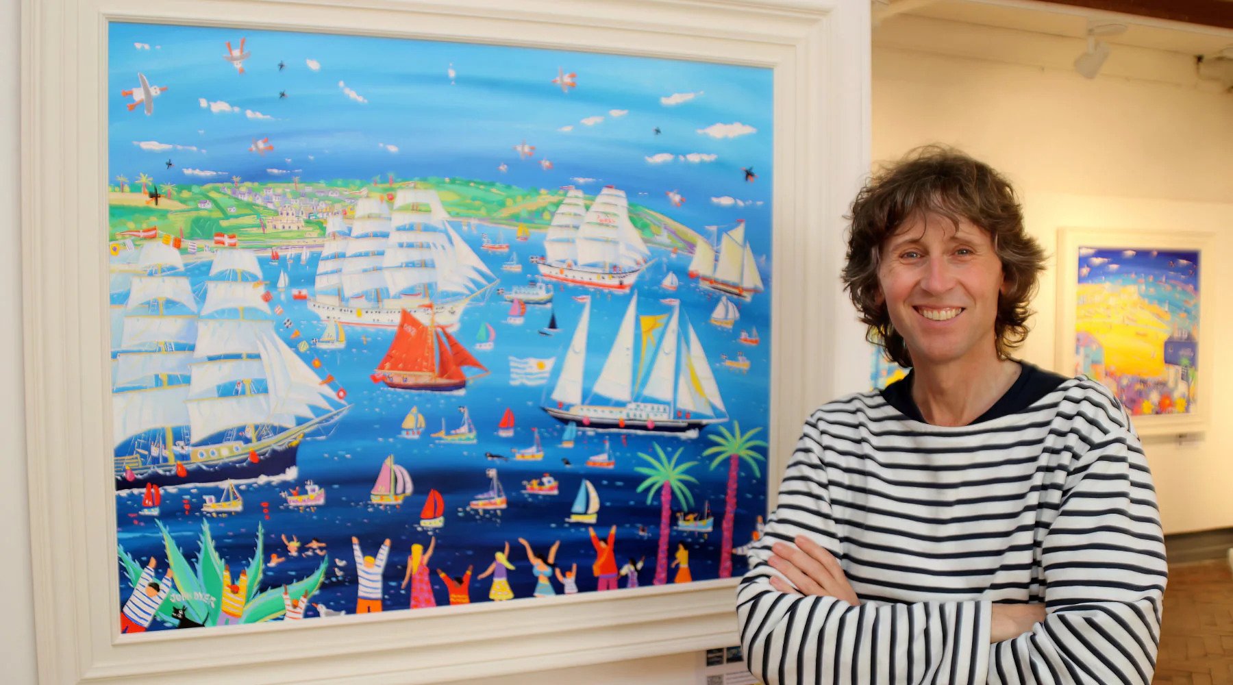A photo of John Dyer, Artist in Residence for Tall Ships Races 2008, 2014 and 2023, at the unveiling of the official painting for 2023 ‘Tall Ships and Small Ships’. John has curly brown hair and is wearing a white and navy striped long-sleeve shirt. He is standing in front of his large framed original painting, which features a colourful scene of tall ships in Falmouth Bay.