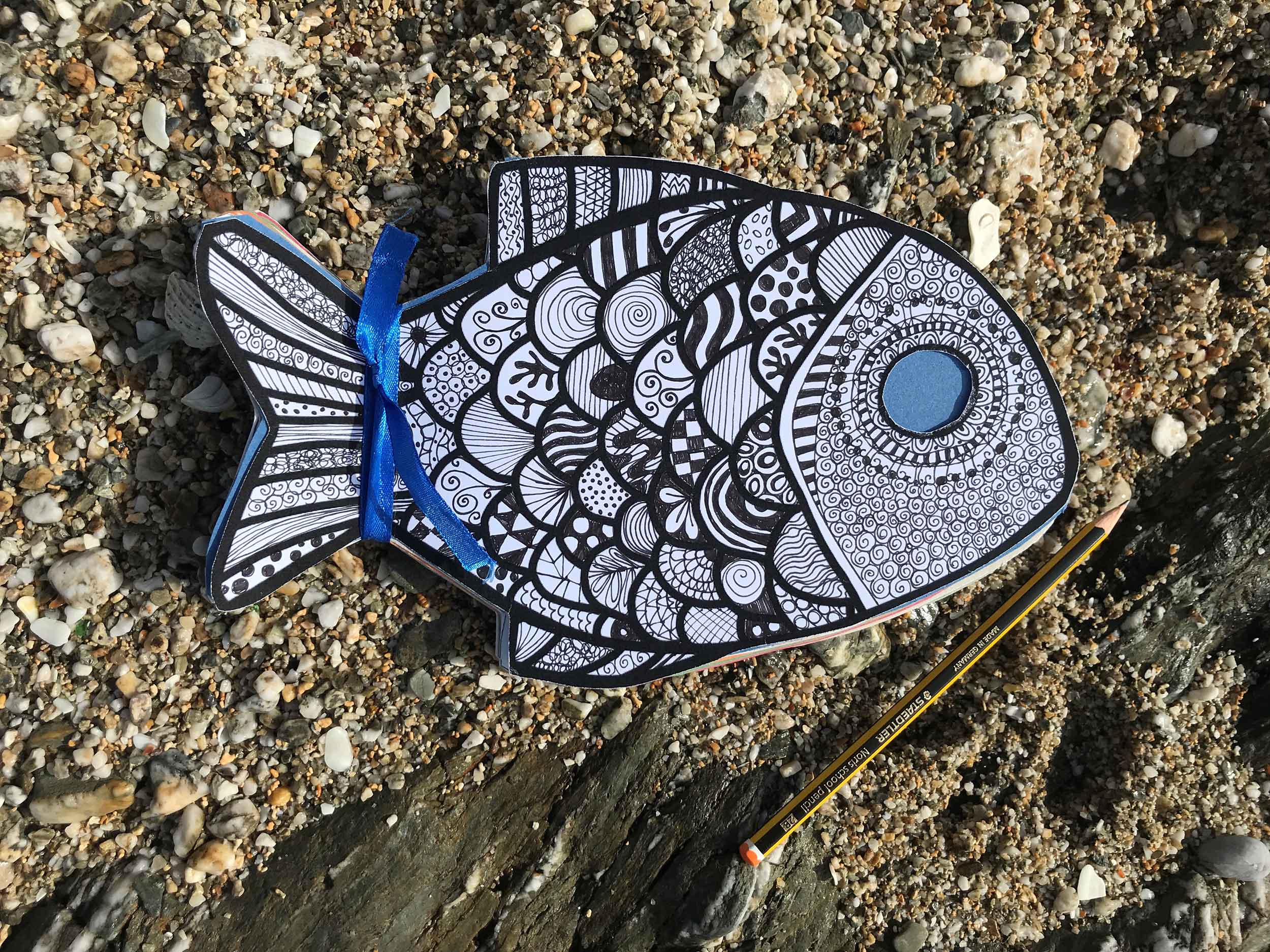 A photo of a fish-shaped colouring in template and a pencil laid out on sand,