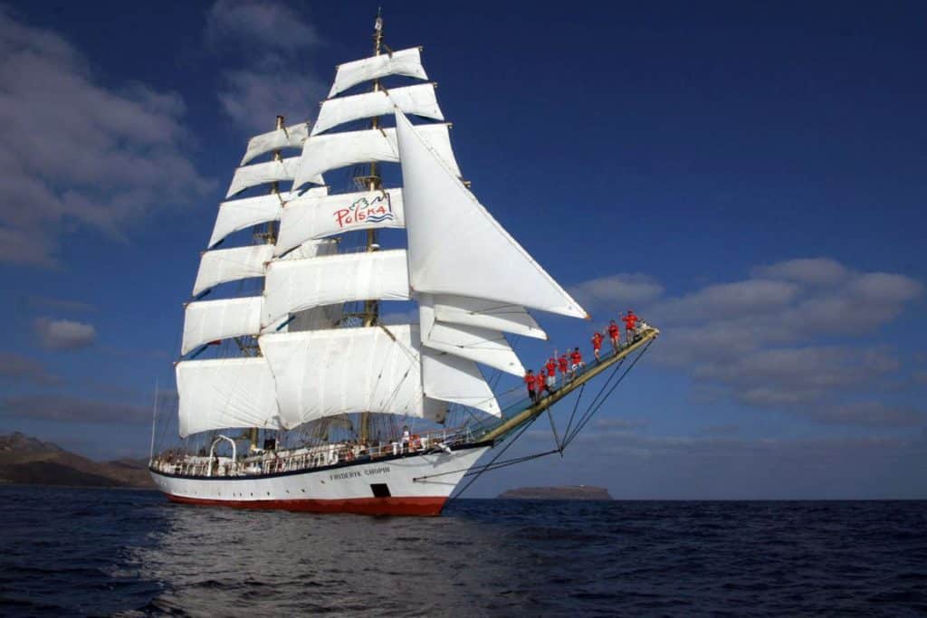 A photo of the Polish tall ship Fryderyk Chopin. The large sailing ship has a red and white hull and white sails. The crew are waving, with some standing on the bowsprit. Pictured on a sunny day,