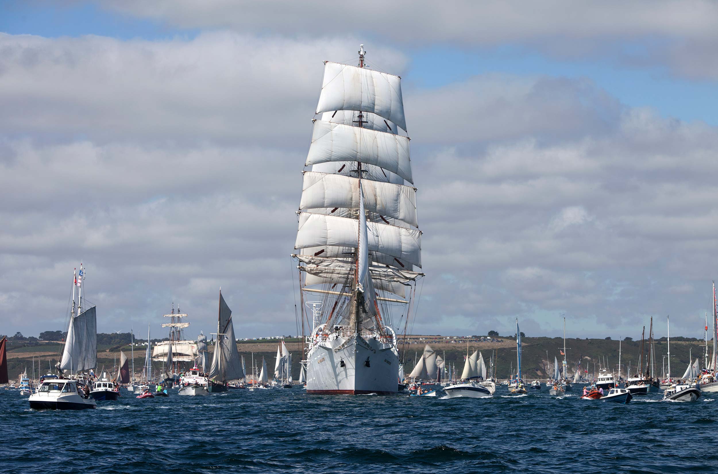 Photo of dozens of ships and boats of different kinds sailing across Falmouth harbour. In the centre is a big sailing ship.