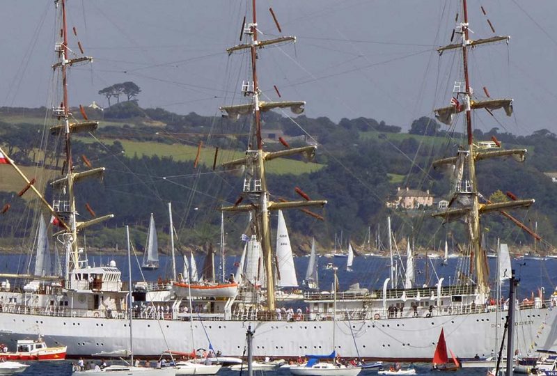 A photo of the Polish Tall Ship Dar Mlodziezy in Falmouth Bay as part of 2014's Parade of Sail. The large white ship is surrounded by smaller sailing vessels. The shot frames Dar Mlodziezy on the water in the centre, with rolling green hills in the background.