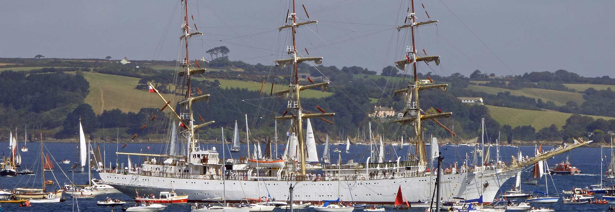 A photo of the Polish Tall Ship Dar Mlodziezy in Falmouth Bay as part of 2014's Parade of Sail. The large white ship is surrounded by smaller sailing vessels. The shot frames Dar Mlodziezy on the water in the centre, with rolling green hills in the background.