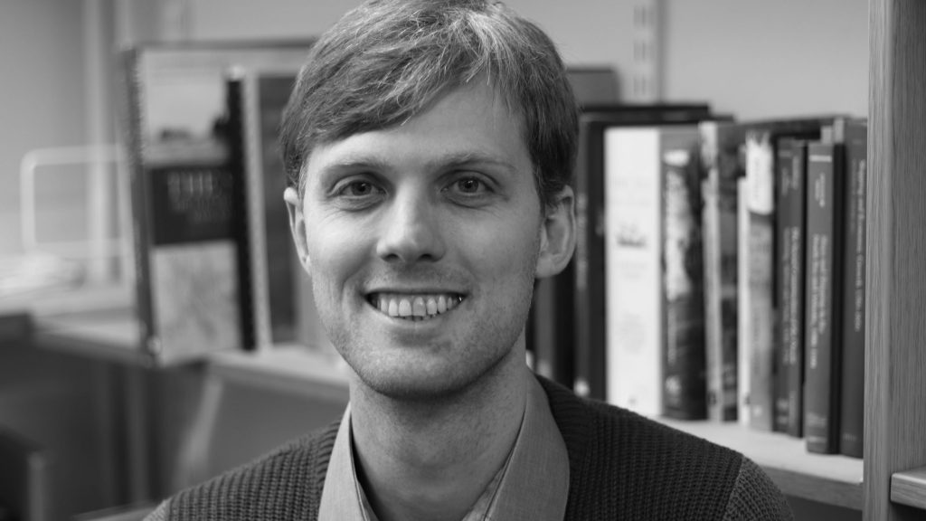 A black and white profile photo of Dr Richard Blakemore. He is smiling at the camera and standing in front of a bookcase.