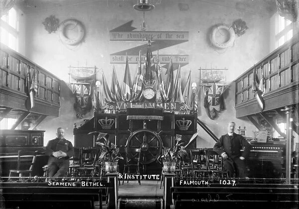 Interior of the Seamen’s Bethel and Institute, founded by the British and Foreign Sailors Society in 1849. Two men are pictured seated by a piano and an organ. There is a pulpit behind them backed by flags and a clock topped with a crown. Life rings from the ship Mohegan of Hull are hung on the wall.