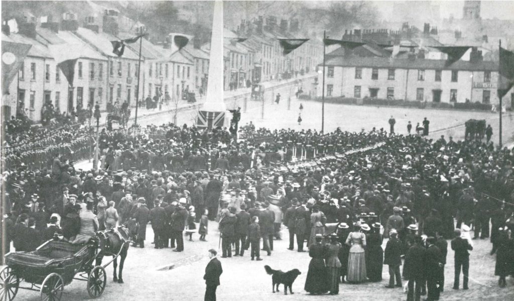 A black and white photo of the unveiling of the Packet Memorial, 19 November 1898. A large gathering of people, all wearing black, are stood around the memorial, which has a large union flag draped over the side of it.