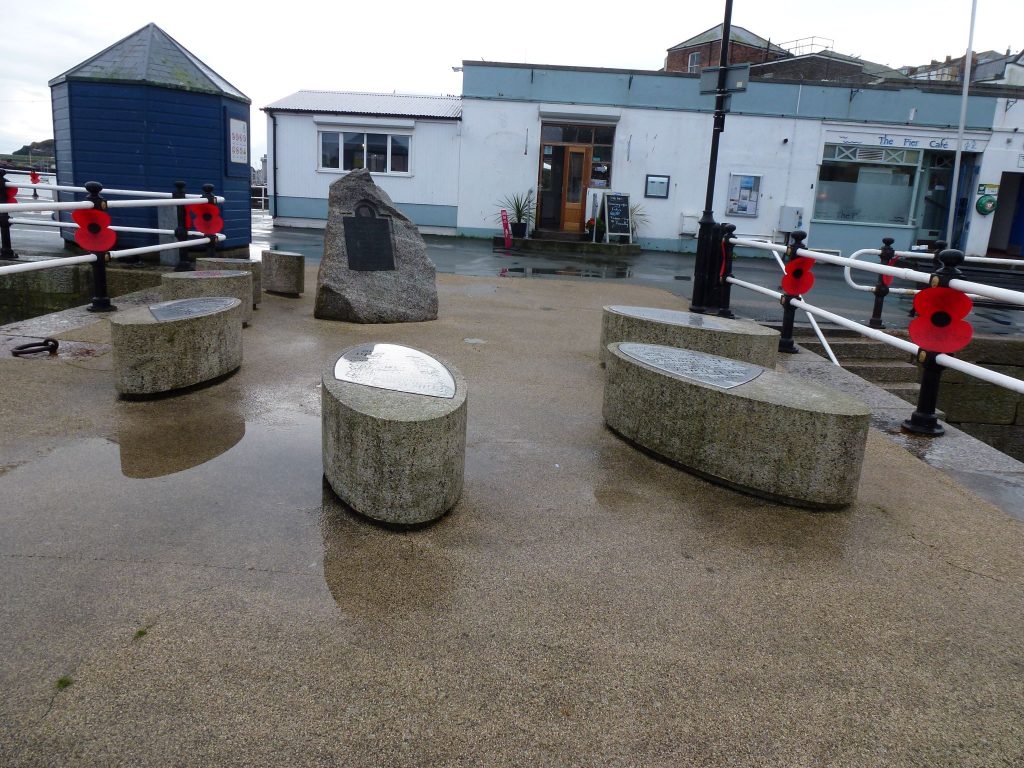 A photo of the St Nazaire Memorial on Prince of Wales Pier; a granite boulder and plaque surrounded by seven oval granite blocks.