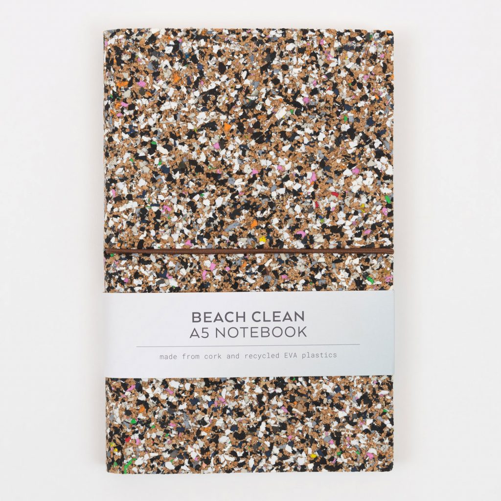 Front of the Beach Clean Cork Notebook. The cover is speckled with lots of different colours. The photo shows the paper packaging going around the notebook.