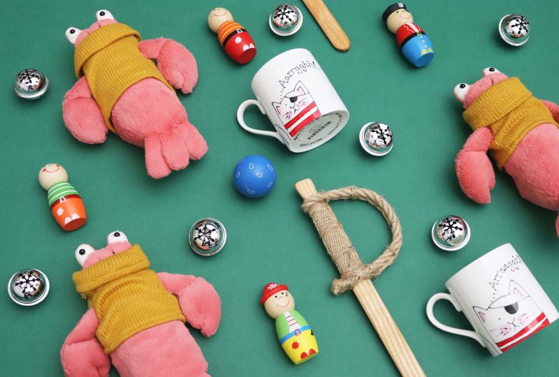 A collection of children's Christmas gifts arranged on a green background. They include a plush lobster, wooden sword, and a pirate cat mug. There are silver baubles scattered around for decoration.