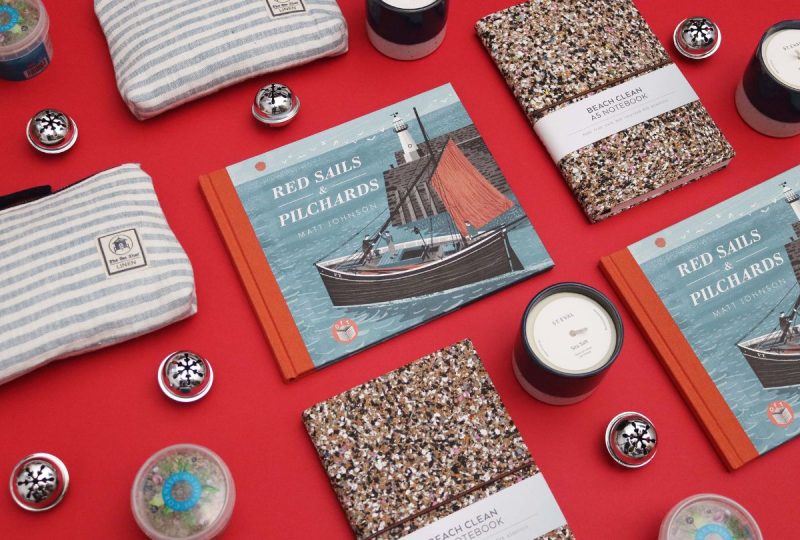 A selection of Cornish Christmas gifts arranged on a red background interspersed with silver bauble decorations. The gifts include a blue and white stripe linen makeup bag, a scented St Eval candle, and an illustration book with a fishing boat on the cover.