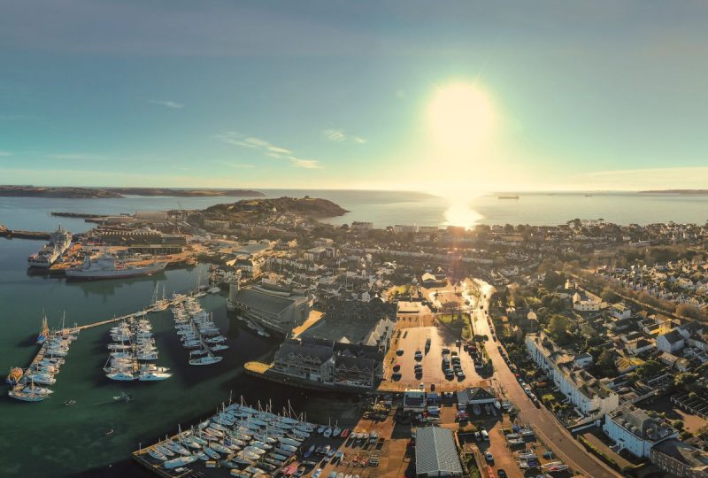 An aerial drone photo of Falmouth overlooking the harbour, National Maritime Museum, and across to Pendennis Headland and out to sea. The photo is taken on a sunny day.