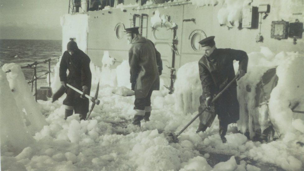 Three sailors with a pick axe and a broom removing ice from a ship.