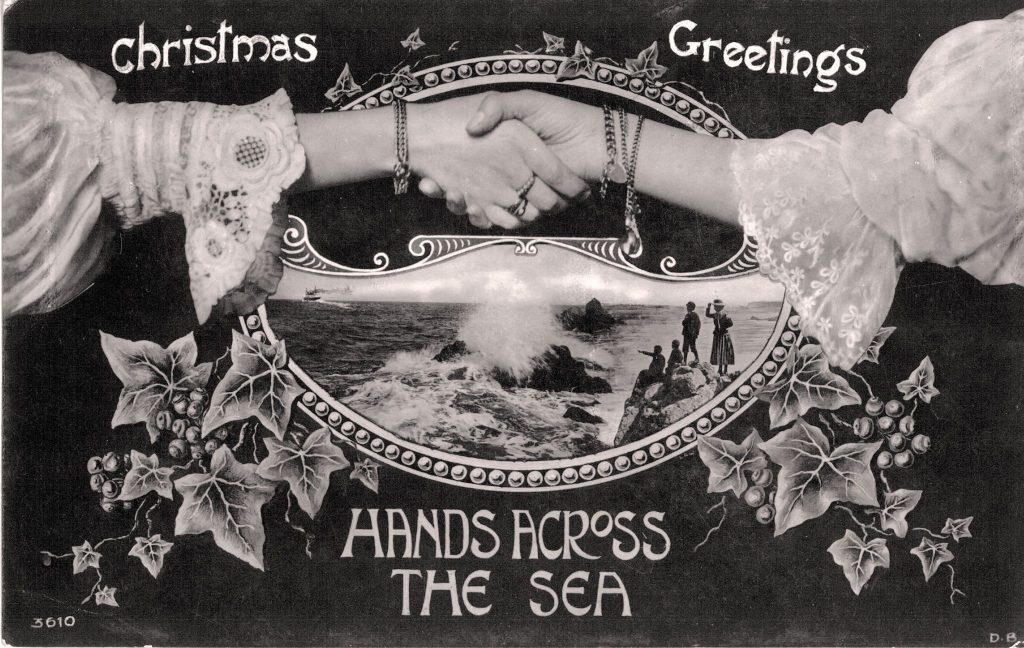 A black and white scan of a festive postcard with the words 'hands across the sea' on it. Two people are shaking hands, underneath their hands is a picture of waves crashing against rocks and people pointing at the sea.