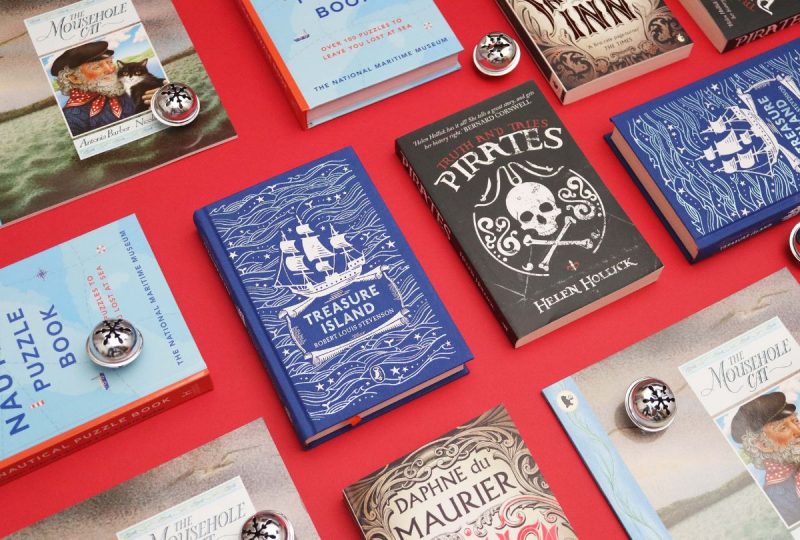 A collection of books arranged on a red surface, interspersed with silver baubles. The books include Jamaica Inn, Treasure Island, Pirates: Truth and Tales, The Nautical Puzzle Book, and The Mousehole Cat.