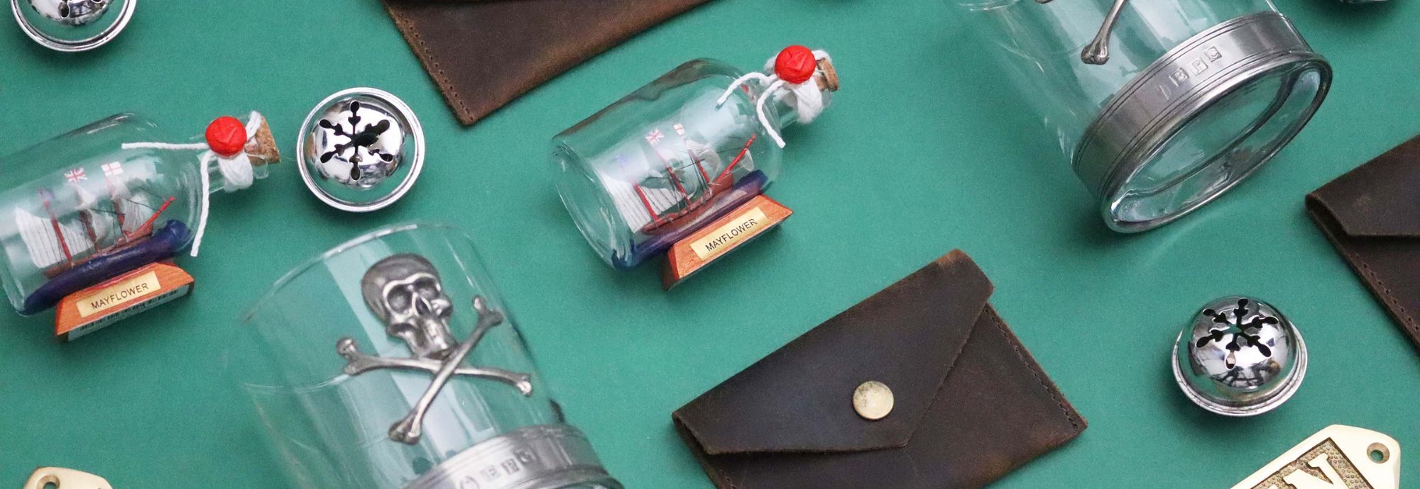 A collection of maritime-themed Christmas gifts arranged on a green table. The gifts include a skull and crossbones whisky tumbler, a leather coin pouch, a brass bottle opener with the text 'Captain' on it, and a small ship in a bottle. There are also silver bauble decorations dotted about.