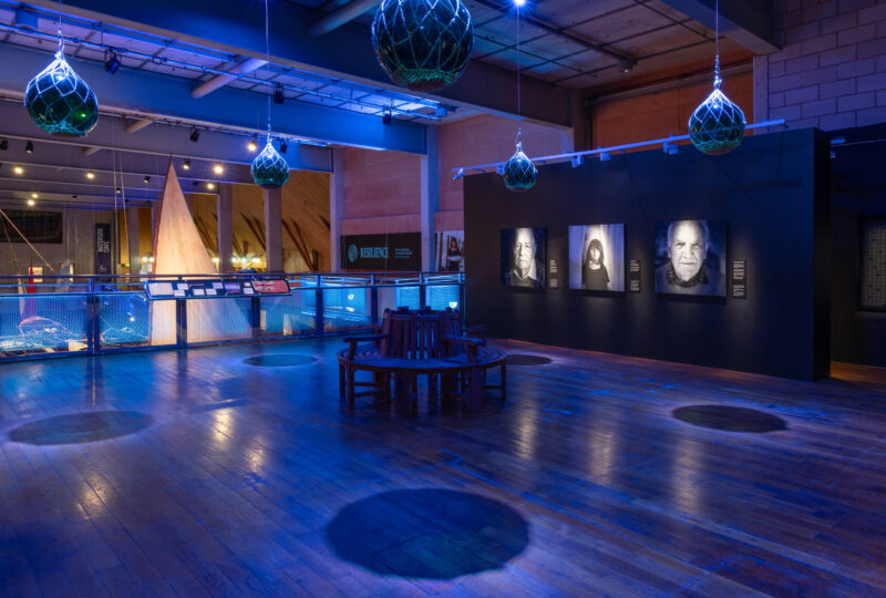 A photograph showing an exhibition space with black walls and a line of large black and white portraits. In the middle of the space a circular bench sits under suspended glass buoys which are casting shadows on the wooden floor.