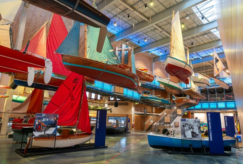 A photo of the Boat Hall at National Maritime Museum Cornwall. Some boats hang from the ceiling, others are positioned on the ground.