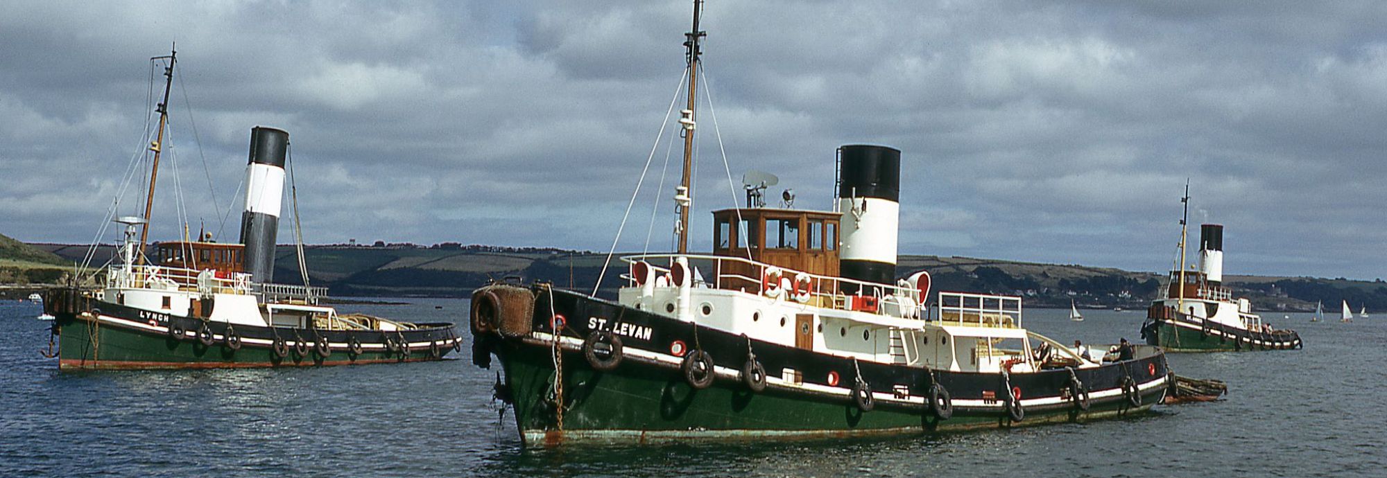 Three steam tugs sailing in Falmouth Harbour. The tugs are green, black, and white, with large funnels. It's an overcast day.