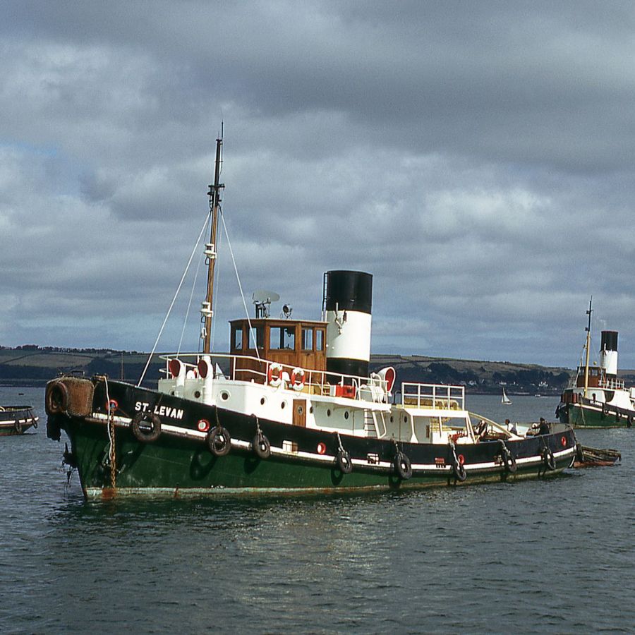 Three steam tugs sailing in Falmouth Harbour. The tugs are green, black, and white, with large funnels. It's an overcast day.