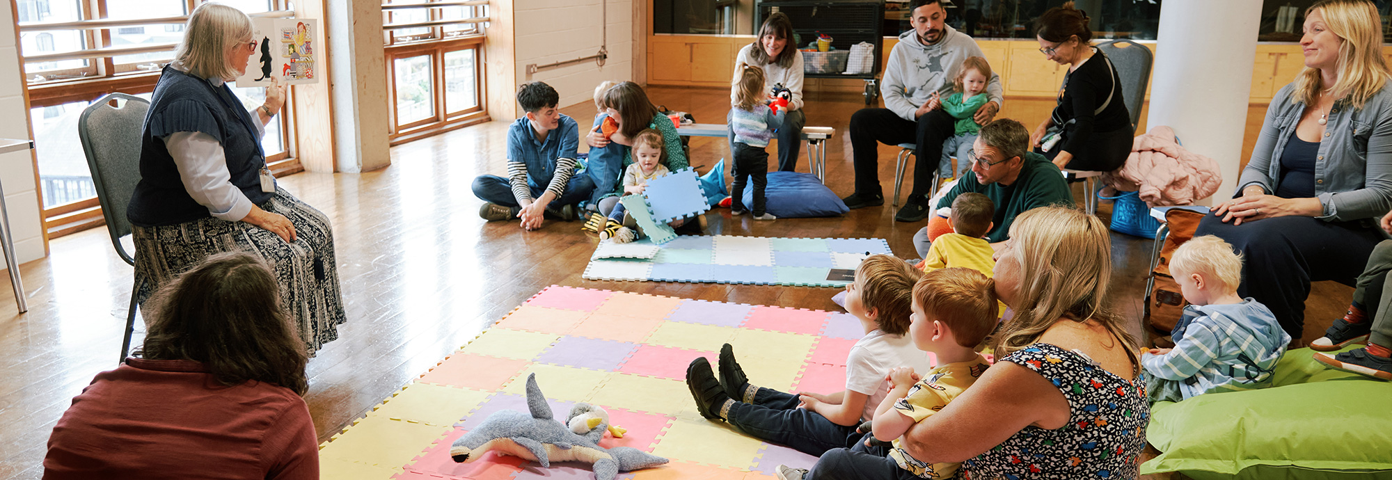 A photo of a lady reading a story to a group of adults and children.