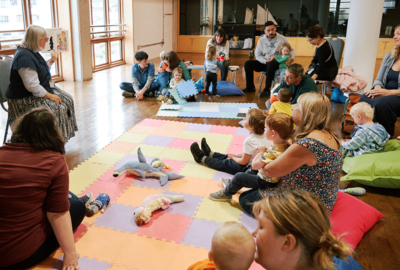 A photo of a lady reading a story to a group of adults and children.