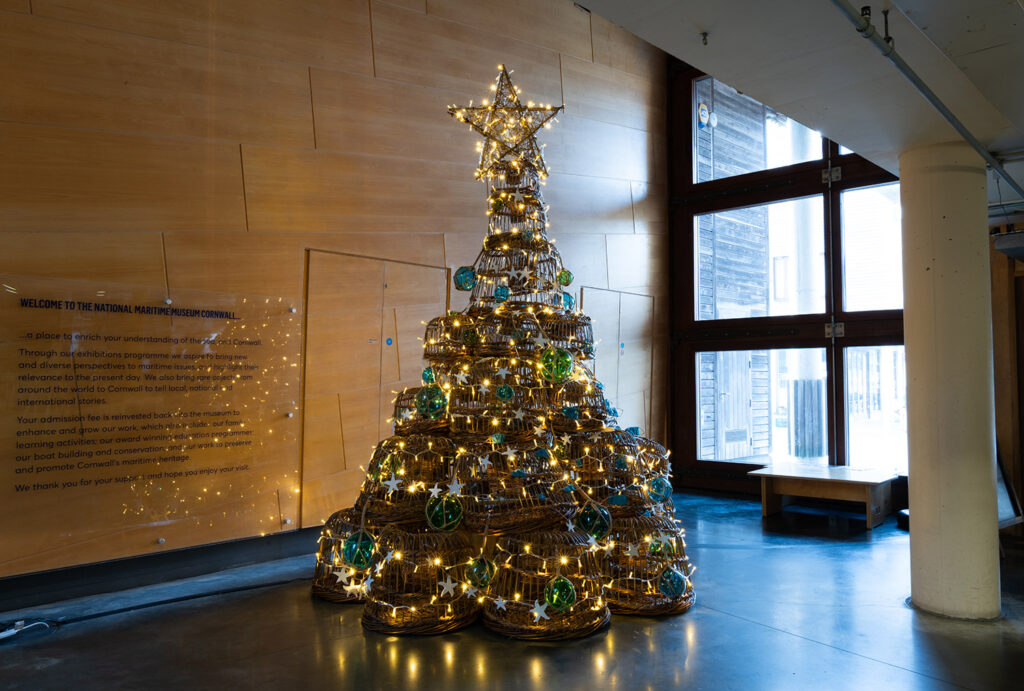 A photo of traditional willow lobster pots stacked on top of each other in a shape of a christmas tree. The tree is decorated with glass bouys and fairy lights.