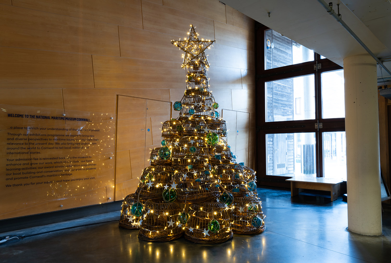 A photo of traditional willow lobster pots stacked on top of each other in a shape of a christmas tree. The tree is decorated with glass bouys and fairy lights.