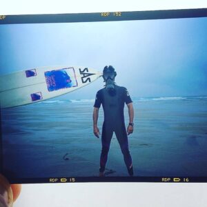 A person in a short sleeve wetsuit wears a gas mask and is standing in front of the ocean, from the side of the image a surfboard is held horizontally pointing towards them.