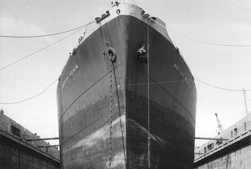 A black and white photograph of a large ship in a dry dock, a line of workers stand underneath the ship.