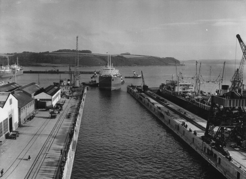 A black and white photograph of a large Tanker entering the dry dock atFalmouth Docks. Employees of Falmouth Docks can be seen lining the sides of the dock to watch.