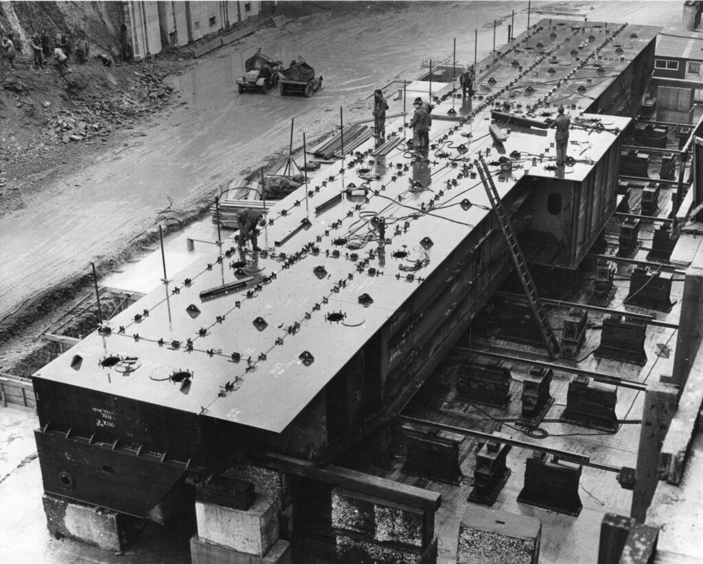 A black and white photograph showing a large section of the Dock Gate being assembled in the dry Dock.