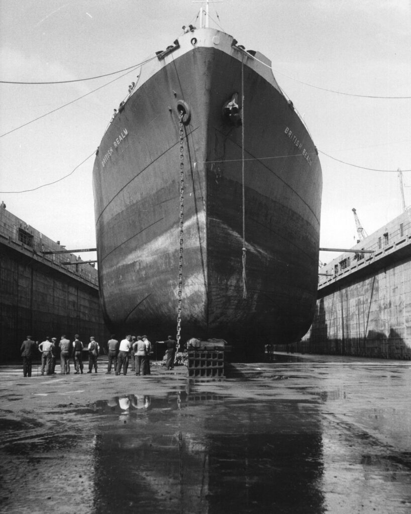 A black and white photograph taken from inside the dry dock showing the large tanker British Realm in the background, with a group of dock workers in the foreground lined up.