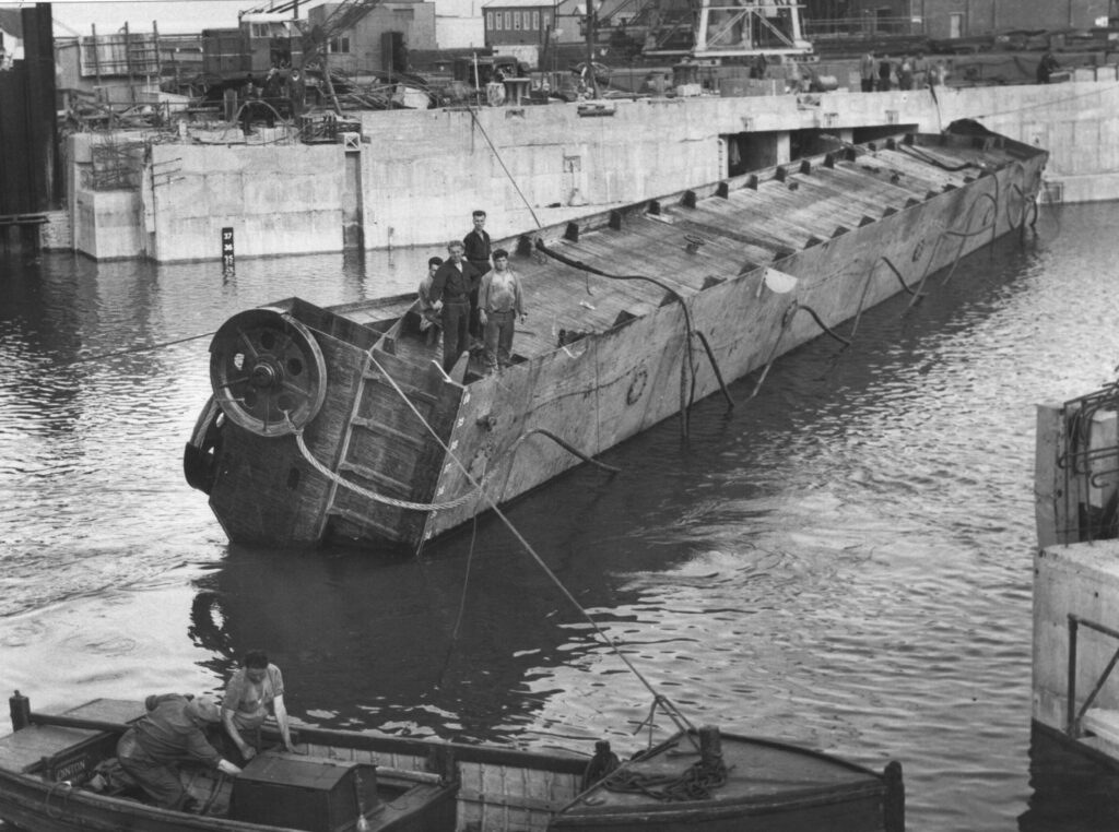 A black and white photograph showing the Dock Gate being floated to position, with four men standing on the floating structure.