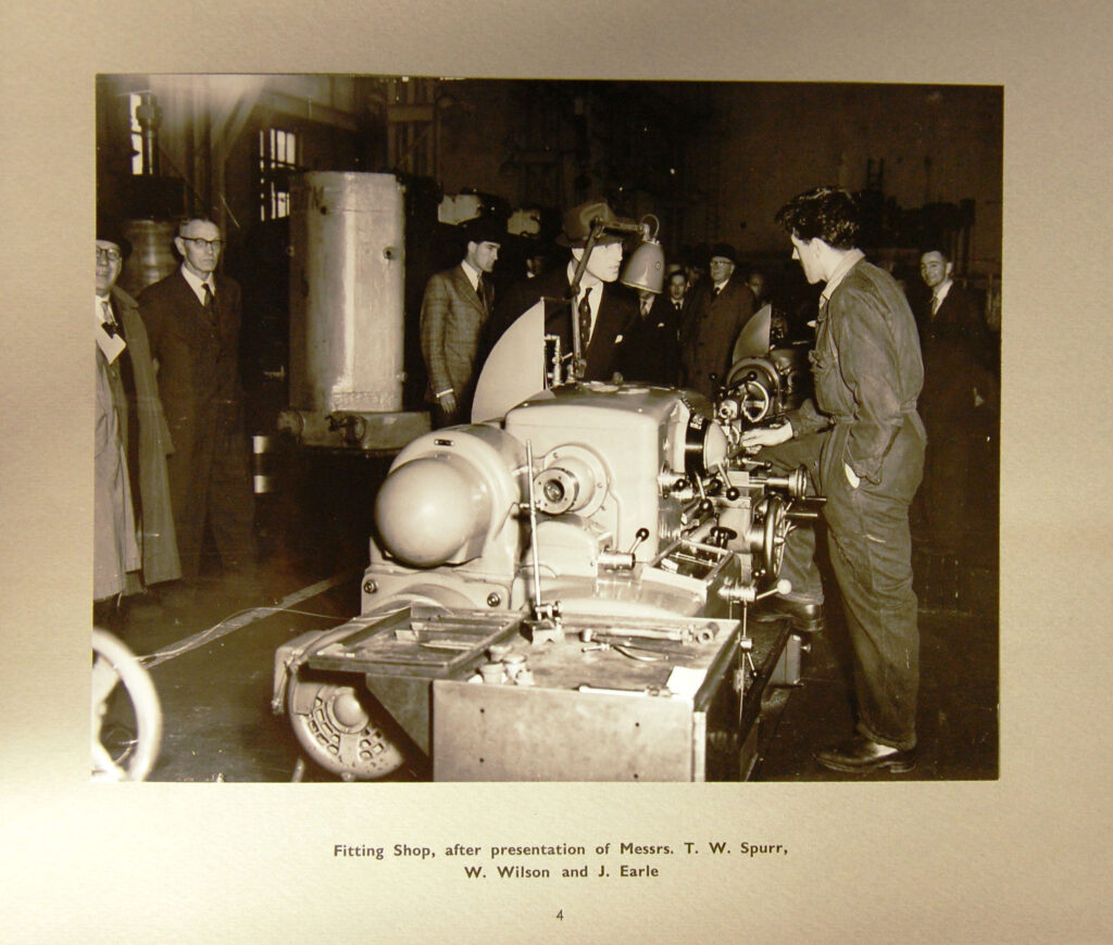 A Black and White Photograph showing a group of people gathered around equipment in the fitting shop, Queen Elizabeth Dock, Falmouth.