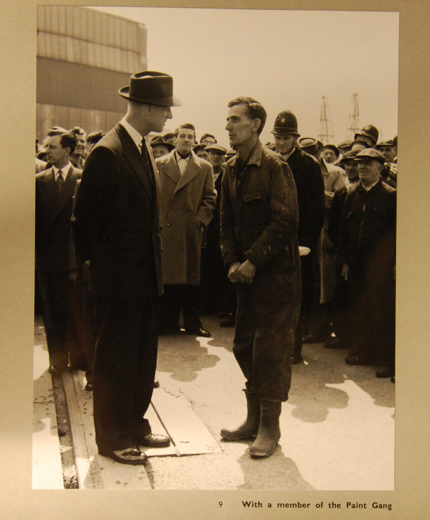 A Black and White Photograph showing one smartly dressed man talking to a man wearing overalls and boots, surrounded by a group of onlookers.