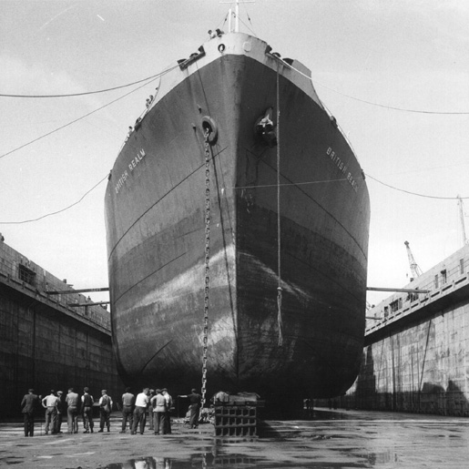 A black and white photograph of a large ship in a dry dock, a line of workers stand underneath the ship.