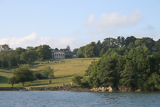 A photograph showing a river in the foreground with a large manor house in the background on the top of a hill surrounded by trees.