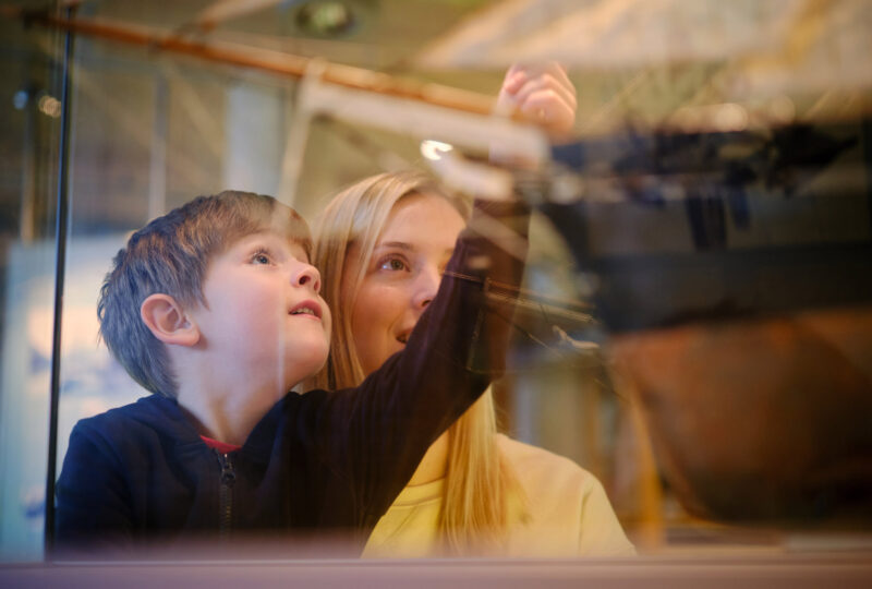 A young boy points to a model ship in a glass cabinet while a lady standing behind him also looks at the ship.