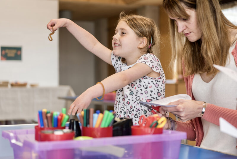 A girl lifts her arm out holding a crayon, there are pots of coloured pencils and pens in front of her and a woman using scissors to cut paper.