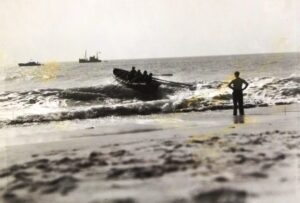 A black and white photograph showing a man with his hands on his hips looking out to a boat being rowed away from the shore.