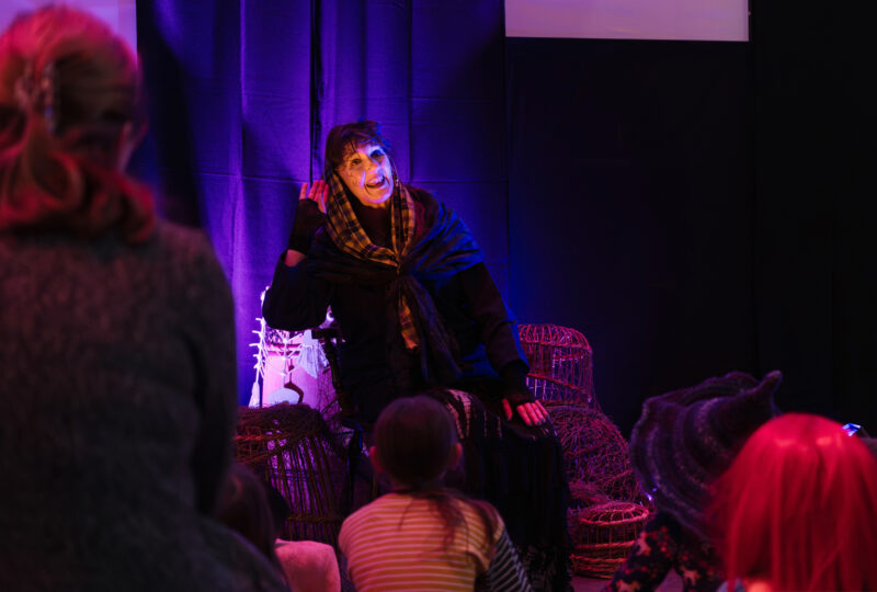 A photograph of a woman wearing a tartan scarf around her head and a black shawl, holding one hand up to her ear while telling stories to an audience of children.