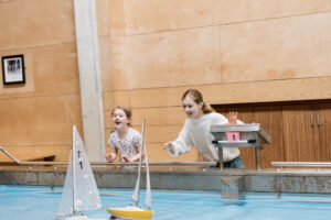Two young girls laugh and point at model boats floating across an indoor pool.