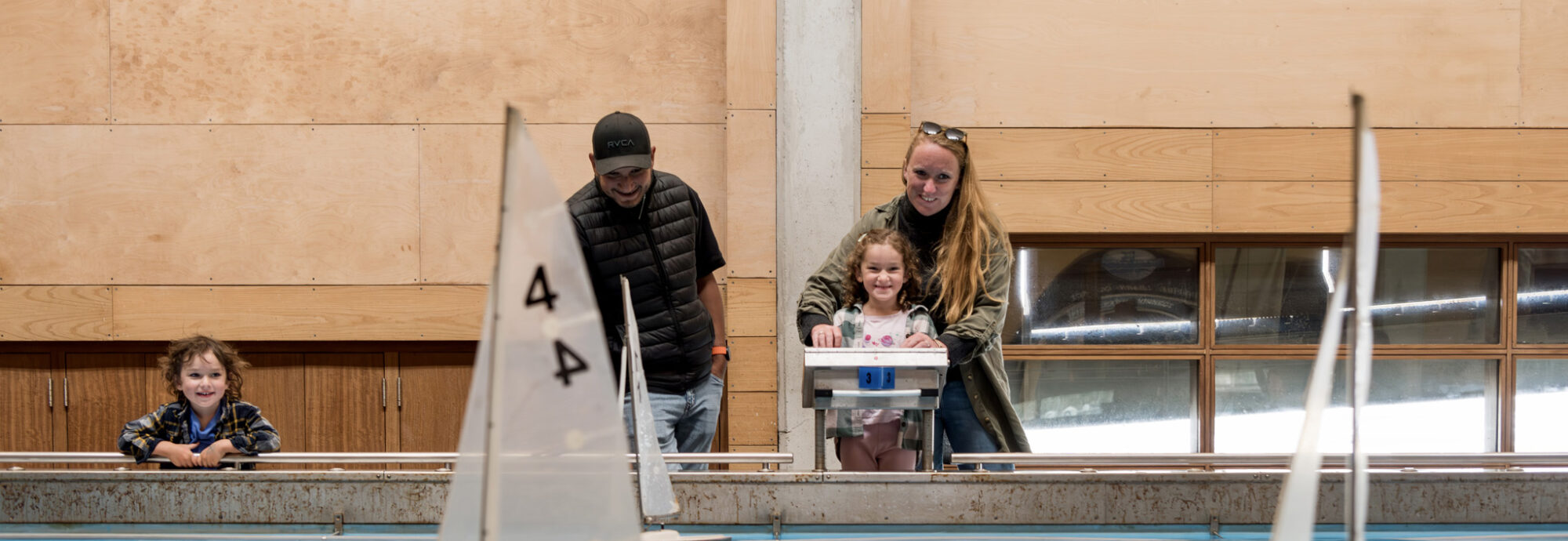 A family of adults and children stand at the side of an indoor pool watching model boats float across the surface.