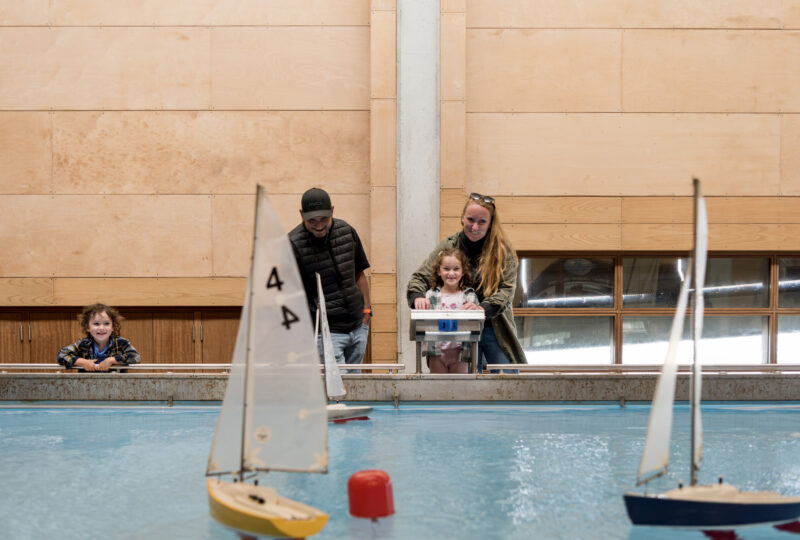 A family of adults and children stand at the side of an indoor pool watching model boats float across the surface.